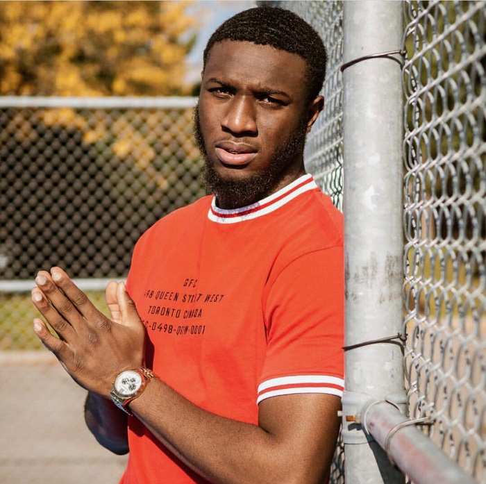 AmanLikeJoseph leans against a chain link fence