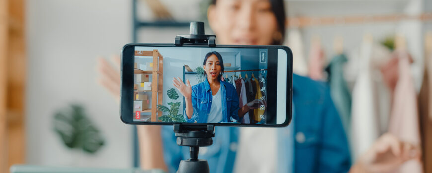 Young Asia lady fashion designer using mobile phone receiving purchase order and showing clothes recording video live streaming online at shop.